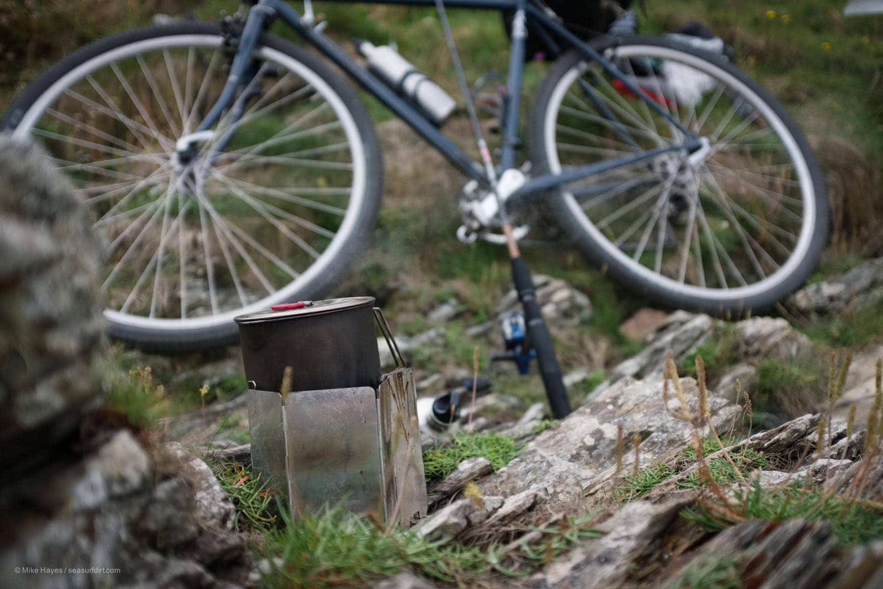 camping stove making tea with a bicycle and fishing rod in the background